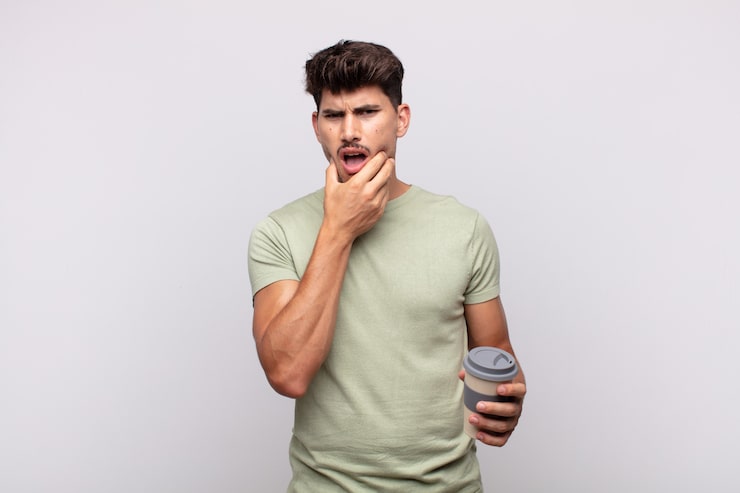 Young man with a coffee with mouth and eyes wide open and hand on chin, feeling unpleasantly shocked, saying what or wow